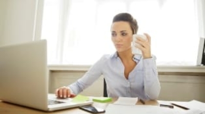 Woman working on a laptop at her office desk while holding a smartphone