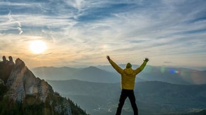 Hiker in a yellow jacket standing on a mountain peak with arms outstretched at sunset