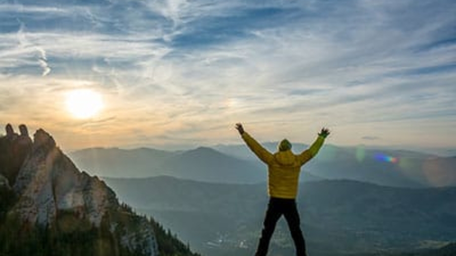 Hiker in a yellow jacket standing on a mountain peak with arms outstretched at sunset