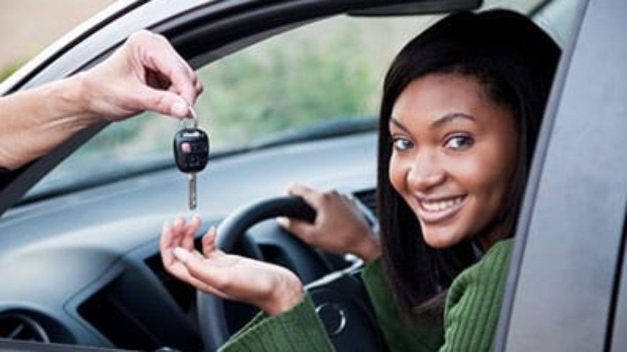 Young woman leaning against her car door smiling and holding up car keys on a city street