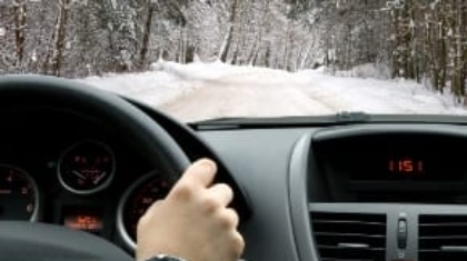 Driver POV behind the steering wheel navigating a vehicle along a snow-covered road lined with pine trees