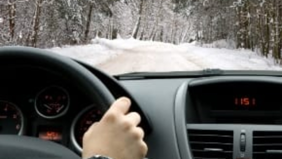 Driver POV behind the steering wheel navigating a vehicle along a snow-covered road lined with pine trees