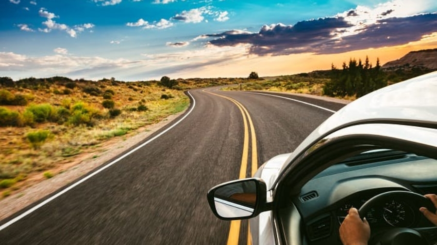 Driver holding the steering wheel while driving along a winding open road under a scenic sunset