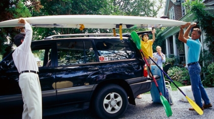 Family loading a paddleboard onto the roof rack of a black SUV parked outside a home