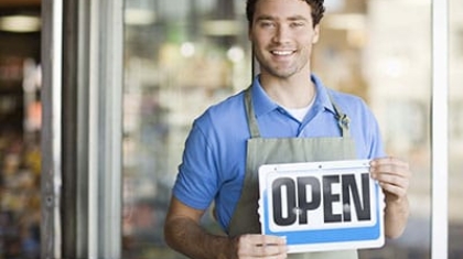 Smiling small business owner wearing an apron holding an open sign at a storefront entrance