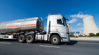 Large silver fuel tanker truck driving along a highway past power plant cooling towers