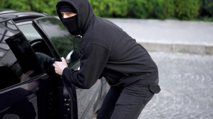 Person wearing a black hoodie and face mask breaking into a parked car door