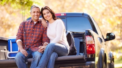 Couple sitting on the open tailgate of a dark pickup truck outdoors holding fishing rods