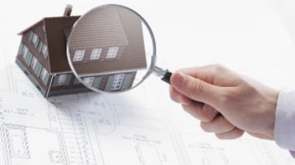 Hand holding a magnifying glass over a small model house on top of architectural blueprints