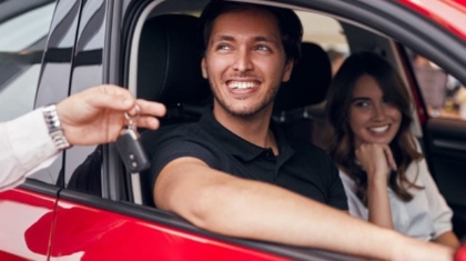 Smiling man behind the wheel receiving keys to a red rental car from an agent