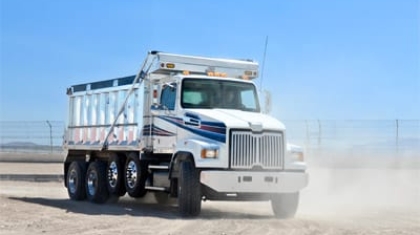 White multi-axle dump truck driving on a dusty work site under a clear blue sky