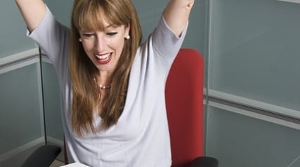 Excited woman sitting at an office desk celebrating with arms raised in victory