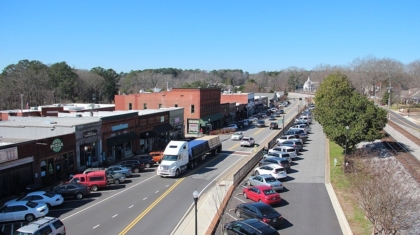 Elevated view of a commercial town street filled with traffic, parked cars, and storefronts along a railway track