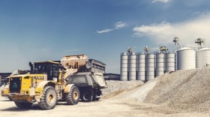Yellow front wheel loader loading materials into a dump truck at an industrial worksite near storage silos