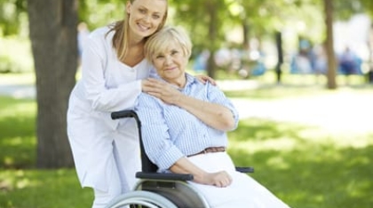 Female caregiver standing behind and comforting an elderly woman sitting in a wheelchair outdoors in a park