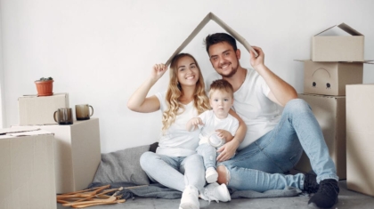 Family holding a cardboard roof cutout over their heads while sitting near moving boxes