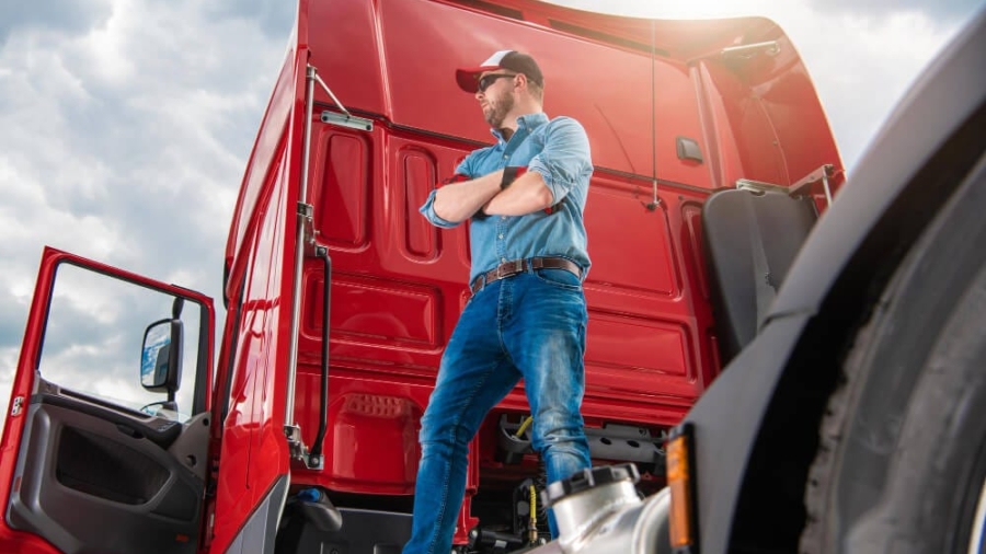 Truck driver wearing a cap standing next to the cabin of a large red semi-truck