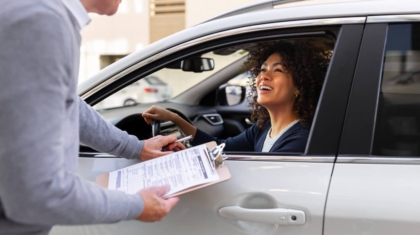 Insurance agent handing a clipboard with paperwork through a car window to a smiling female driver