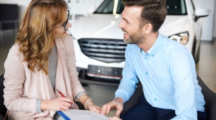 Female insurance agent reviewing paperwork on a clipboard with a smiling young driver sitting in front of a white car