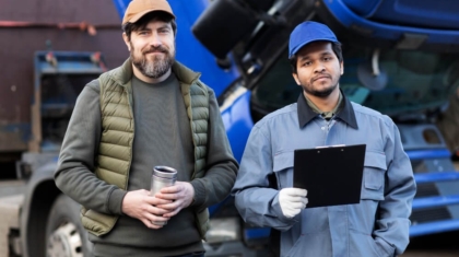 Two truck drivers standing in front of a blue semi-truck, one holding a travel mug and the other holding a clipboard