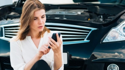 Woman sitting on the ground using her smartphone in front of a car with an open hood