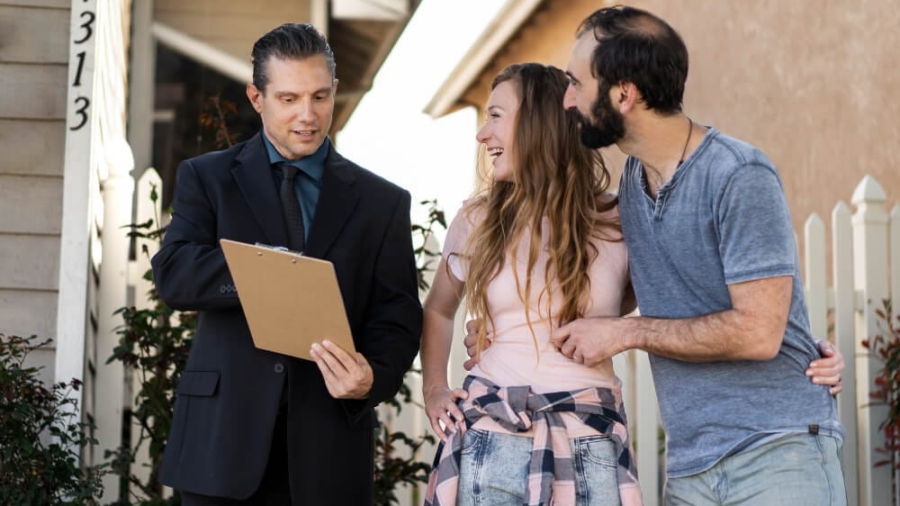Insurance agent holding a clipboard while reviewing a home insurance claim with a young couple outside their house