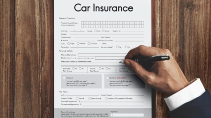 Hand filling out a printed car insurance application form on a clipboard placed on a wooden desk