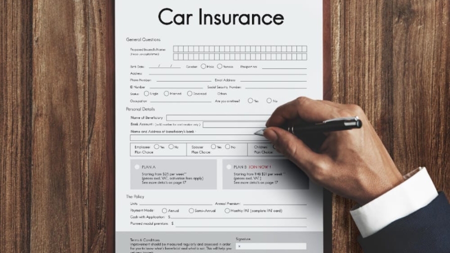 Hand filling out a printed car insurance application form on a clipboard placed on a wooden desk