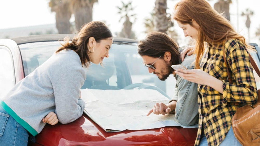 Group of three young adults looking at a paper road map on the hood of a car outdoors
