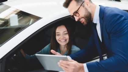 Car salesman in a suit pointing at a digital tablet while showing features to a smiling woman inside a white car