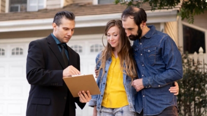 Insurance agent in a suit showing clipboard documents to a couple standing outside a residential home